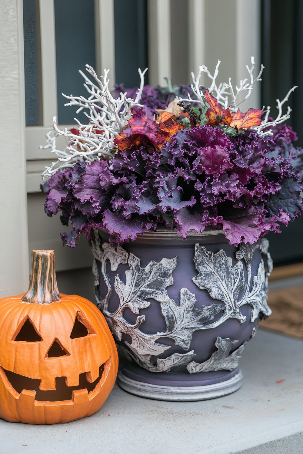 An image depicting autumnal decor on a porch. A carved pumpkin lantern with a cheerful face sits next to a decorative pot filled with vivid purple ornamental kale, white decorative branches, and colorful autumn leaves. The pot has raised leaf designs painted in a weathered grey finish, adding texture and visual appeal to the arrangement. The background features beige-painted wood paneling.