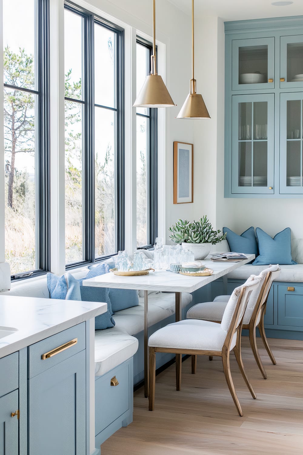 Bright kitchen nook with light blue cabinetry, white countertops, wooden chairs with white cushions, pendant lighting, many windows, and a potted succulent on the table.