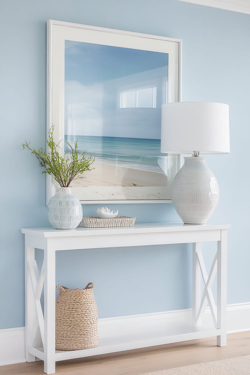 A console table against a pastel blue wall, decorated in a coastal style. The table includes a large framed beach photograph featuring serene blue ocean waves and sandy beach. On the table, there is a light blue ceramic vase with green branches, a round wicker tray holding a white ceramic lotus, and a white textured table lamp with a plain white lampshade. Below the table, a woven basket rests on the lower shelf.