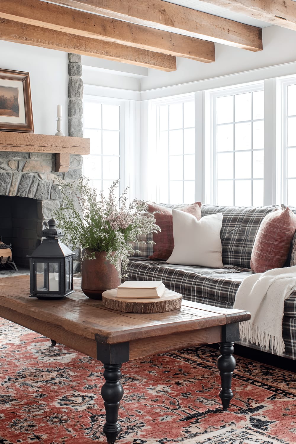 A rustic cabin interior featuring a comfortable vintage plaid sofa strewn with chunky knit blankets. A Persian rug covers the wooden floor while a wooden coffee table graced with antique pieces is the focal point. Large wooden-framed windows bathe the room in natural light. The room's charm is heightened by the presence of reclaimed wooden beams and a traditional stone fireplace.