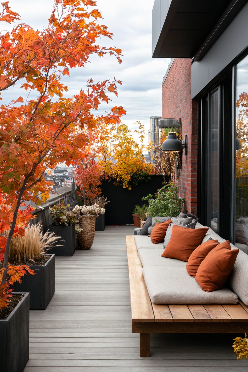 An outdoor terrace features a grey wooden deck with black potted plants and trees showcasing vibrant autumn leaves in shades of red, orange, and yellow. A sleek wooden bench with a beige cushion and orange throw pillows is positioned against a brick wall adorned with a modern black sconce light. In the background, a cityscape with tall buildings is visible under a cloudy sky.