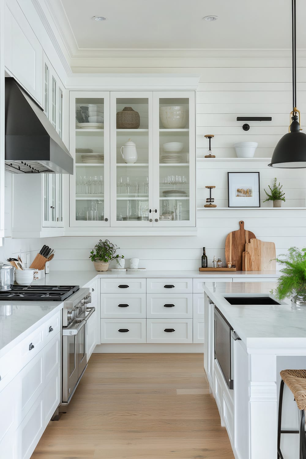 A pristine, modern farmhouse kitchen featuring white cabinetry with black handles, white shiplap walls, light wood flooring, and marble countertops. Open shelves and glass-front cabinets showcase neatly arranged dishware and decor. A black pendant light hangs above a kitchen island with a built-in sink and wicker barstools. Small potted plants and wooden cutting boards add natural elements, while framed artwork brings a touch of personality.