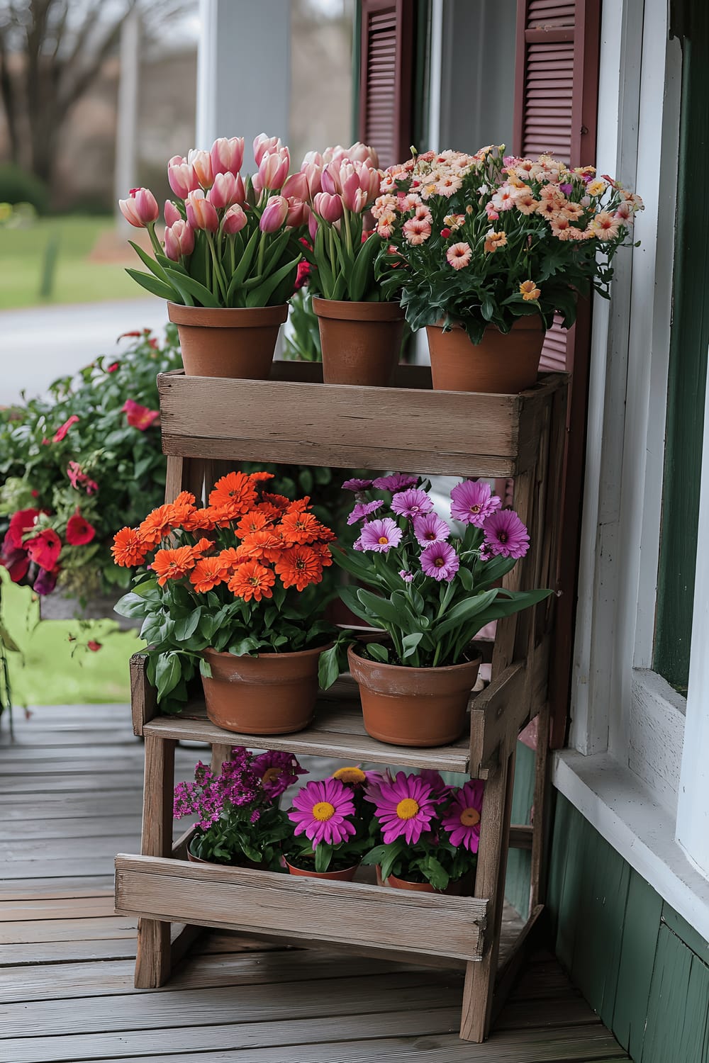 A wooden three-tiered planter stand showcasing a variety of vibrant flowers - tulips, zinnias, and chrysanthemums, arranged in the order of their bloom from spring to autumn. The planter stand is set on a well-maintained garden porch, set against the backdrop of a green outdoor garden.