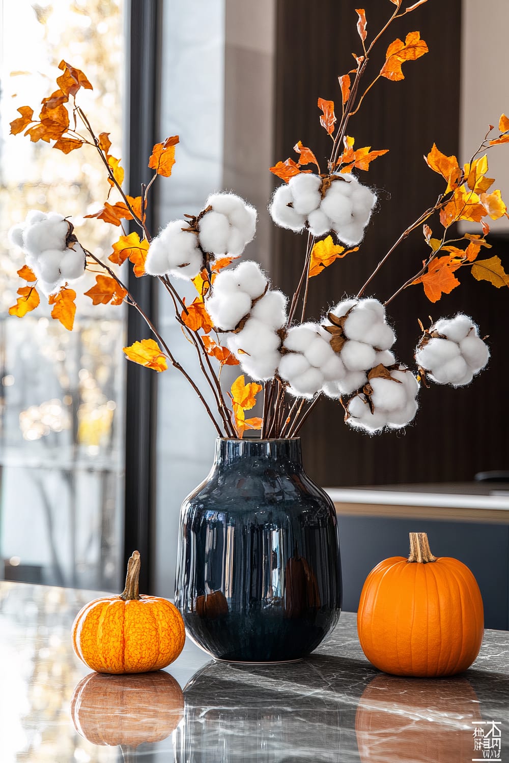 A decorative setup on a polished surface featuring a dark-colored vase filled with cotton branches and orange autumn leaves. Next to the vase, there are two pumpkins, one small and one medium-sized, adding a seasonal touch.