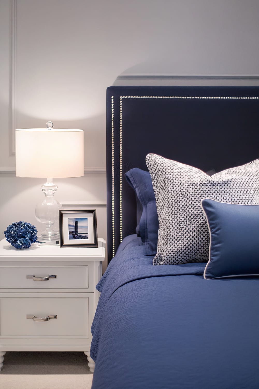 A calming bedroom with a navy blue bed and headboard. A white nightstand features a clear glass lamp with a white shade, a framed photo, and a dark blue decorative object. The bed is made with blue bedding and patterned pillows in white and navy. The room has light grey walls and an organized appearance.