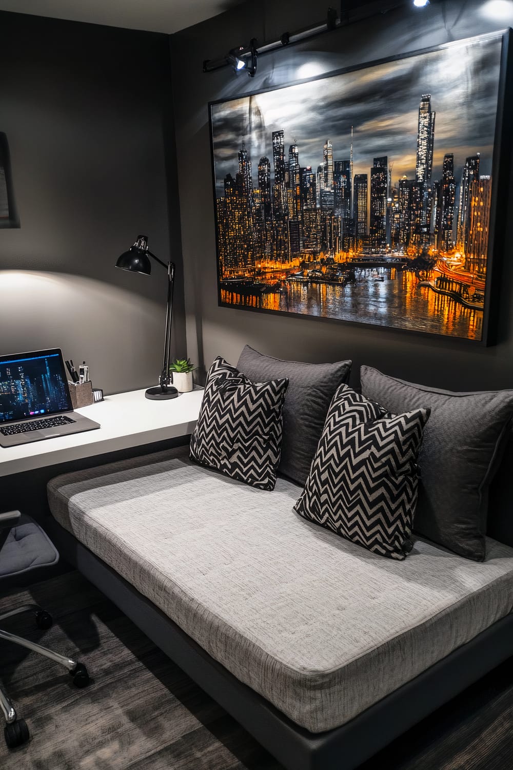 An urban modern room featuring a low-profile grey upholstered bed with black geometric-patterned cushions. A sleek white desk holds a single laptop and a black desk lamp, placed against dark grey walls. A large metallic cityscape art piece dominates the wall above the bed. The room is well-organized, emphasizing contemporary design.