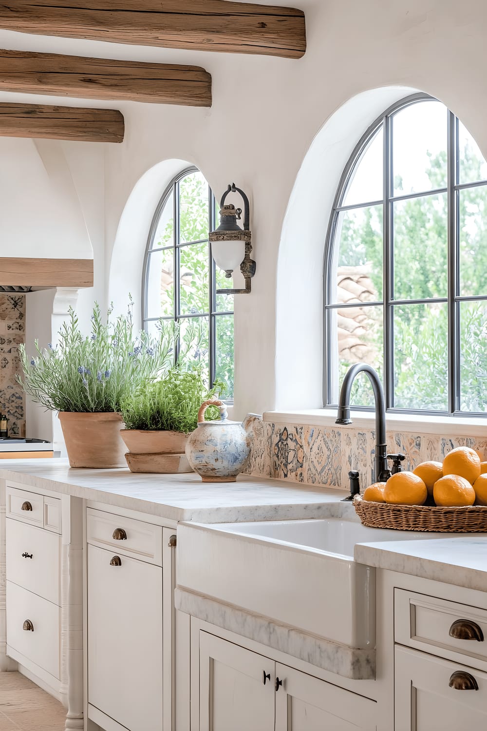 A bright, Mediterranean-style kitchen with creamy white cabinets adorned with blue accents. The room is filled with natural light coming from large arched windows draped with light, airy curtains. The wooden beams and wrought iron light fixtures are highlighted, along with a central island with a marble countertop, holding a vintage olive oil dispenser and a bowl of fresh citrus fruits. The stone countertops and terra cotta tiles add earthy tones, complemented by nautical decors like rope handles, seashell ornaments, and potted lavender plants.