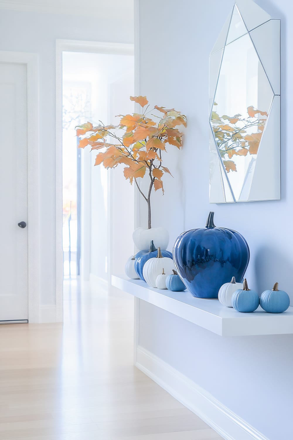 A modern minimalist hallway decorated for Thanksgiving, featuring a sleek white console table with a large potted plant displaying autumn leaves, a minimalist geometric wall mirror, and decorative pumpkins in various shades of blue and white. The space is illuminated by soft natural light, highlighting the clean lines and uncluttered decor.