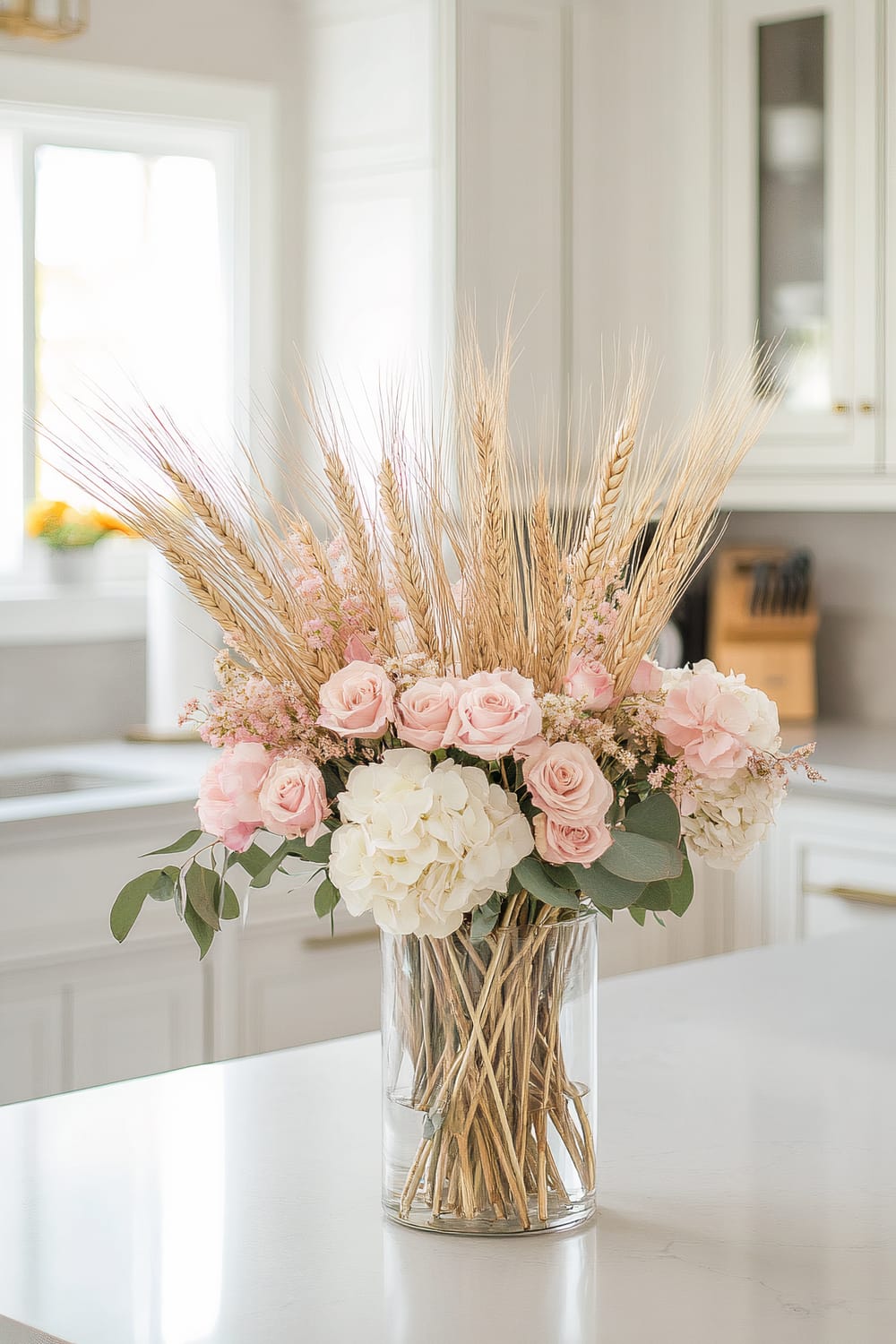 An elegant floral arrangement features a mix of soft pink roses, white hydrangeas, and delicate sprigs of dried wheat in a clear glass vase. The vase is situated on a sleek, white kitchen countertop, with a light-filled window and cabinets visible in the background.