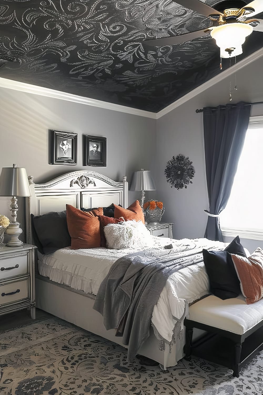 Elegantly decorated bedroom with a dark, intricately patterned ceiling. The bed has an ornate headboard, and it is adorned with white, orange, and black pillows. The room features matching grey nightstands with table lamps, grey walls, and dark blue curtains partially opened to let in natural light. A plush bench is at the foot of the bed, and a patterned rug covers the floor. Wall art and flowers on the nightstand add decorative touches.