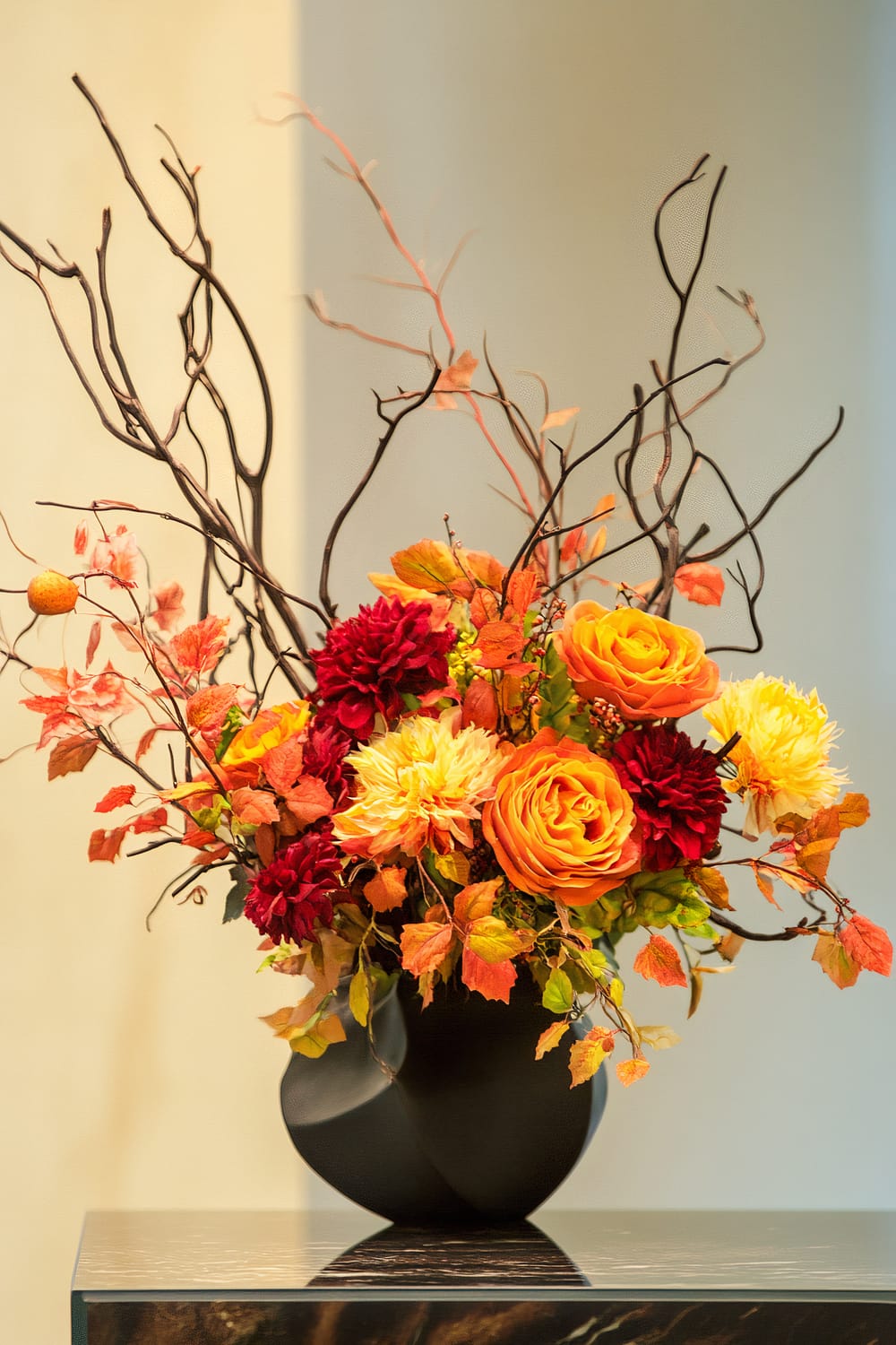 A sophisticated floral arrangement featuring vibrant orange and deep red flowers, along with yellow accents and rustic autumn leaves. The composition is enhanced by dark, twisting branches, all set in a sleek, matte black vase. The arrangement is displayed on a reflective surface, spotlighting the rich colors and textures of the ensemble.