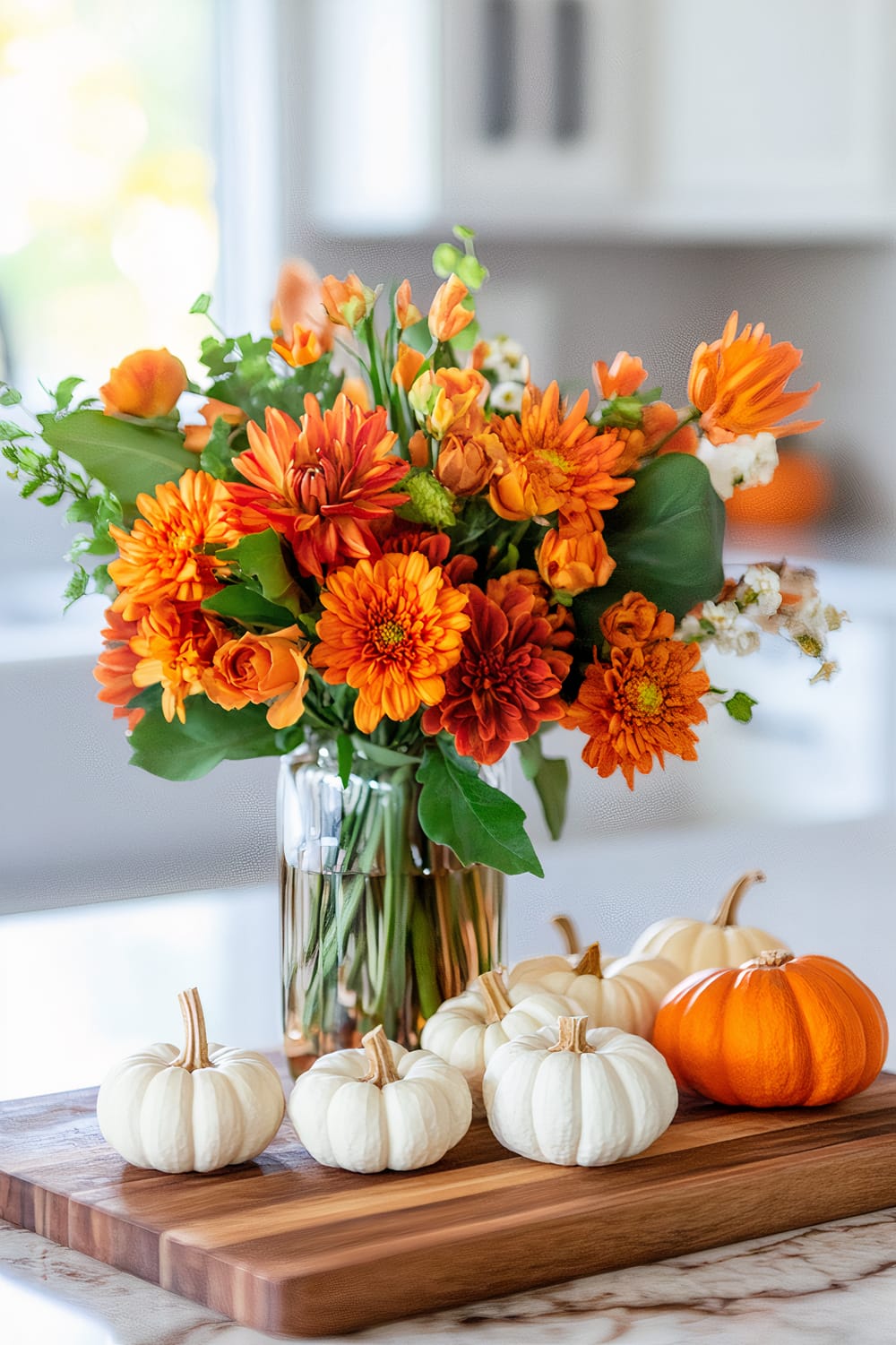 A bouquet of orange and red flowers in a glass vase sits on a kitchen countertop. In front of the vase, there is a wooden cutting board with six white mini pumpkins and one orange mini pumpkin placed on it.