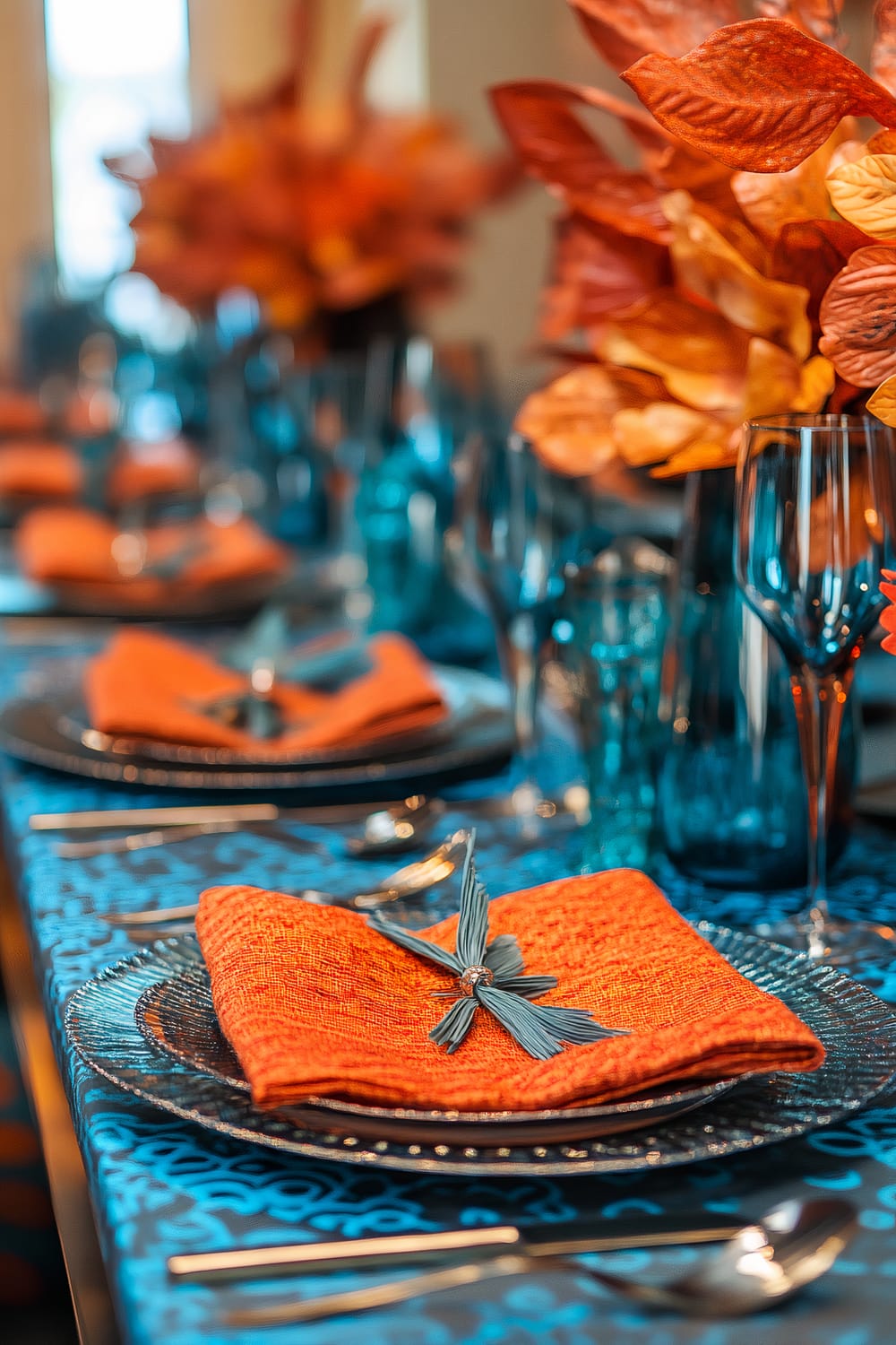 A vibrant table setting featuring bold orange napkins and electric blue glassware. The table is decorated with dynamic centerpiece elements including textured, vibrant orange leaves. The tablecloth has an intricate blue pattern, complementing the color scheme. The overall look is lively and modern.