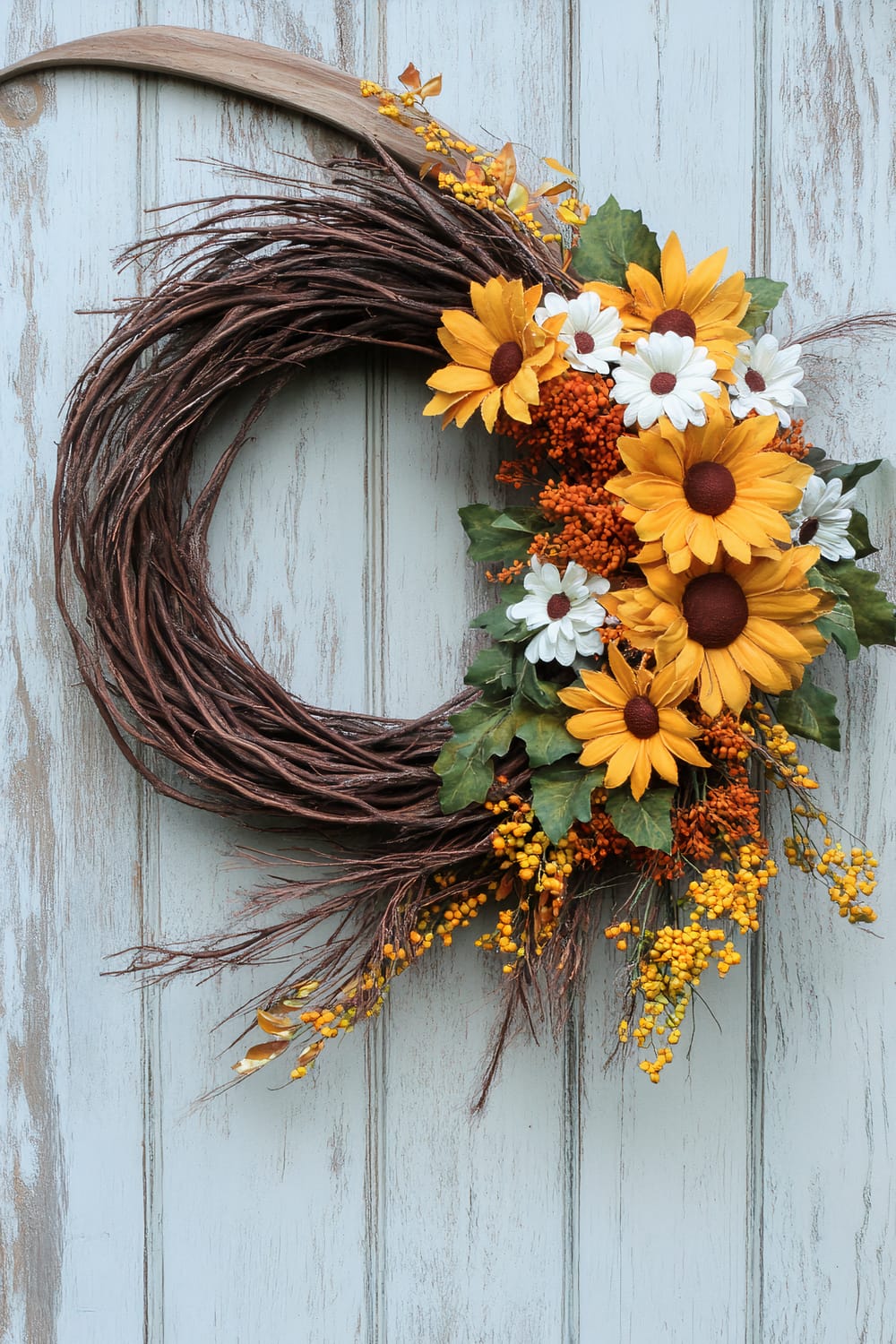 Rustic wreath made from interwoven brown twigs and decorated with artificial yellow sunflowers, white daisies, orange berries, and green leaves. The wreath is displayed on a light blue, distressed wooden background.