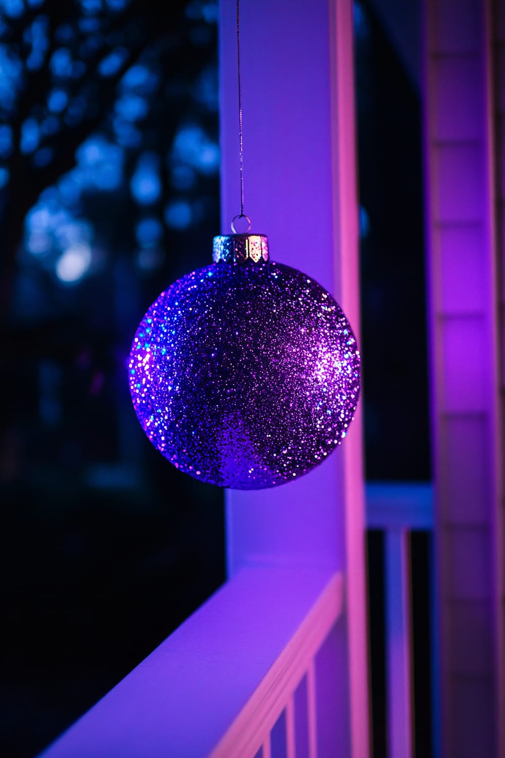 An oversized glittering purple ornament hangs from a sleek white porch railing. The intense purple lighting makes the ornament shimmer against a dark, moody backdrop with hints of trees in the background.