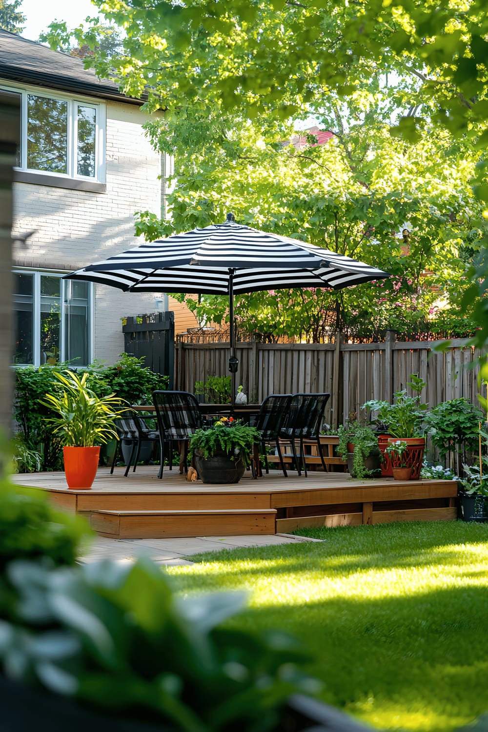 A well-maintained backyard garden featuring a seating area with a table and chairs underneath a bold, black and white-striped umbrella. Vibrant plants filling decorative pots dot the fuel green grass while wooden steps lead from a deck attached to the house to the quaint patio.