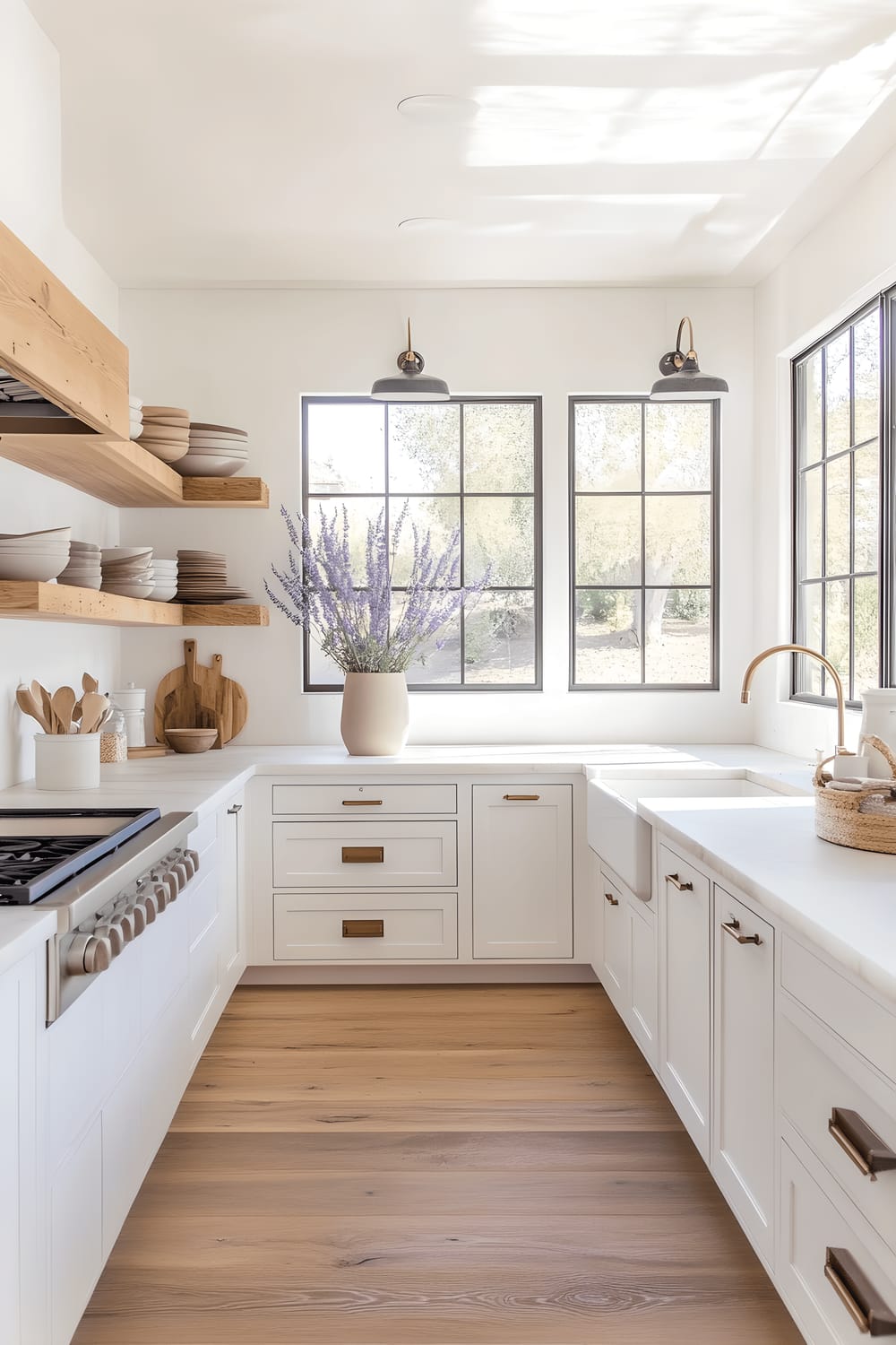 A minimalist kitchen featuring white cabinetry, light oak floating shelves laden with neutral-toned dishware, and a matte vase filled with fresh lavender. A large picture window allows the influx of abundant natural light, illuminating the space and highlighting its sleek elements.