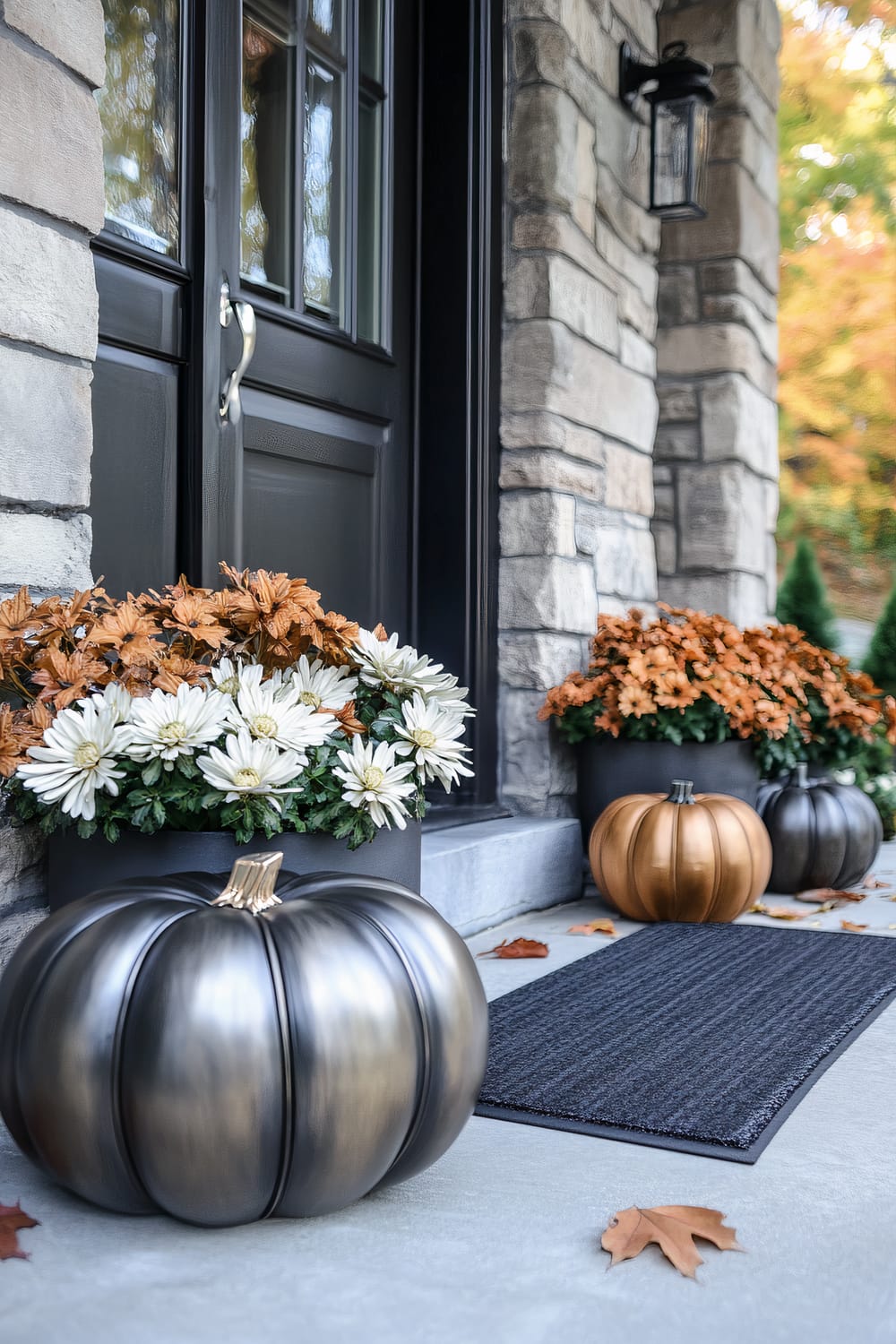 A front porch decor arrangement featuring a sleek black door with a metallic handle, framed by stone walls. Two large planters filled with white and burnt orange chrysanthemums sit on either side of the door. Two metallic pumpkin decorations, one in a gold hue and the other in a bronze tone, are on the porch steps. A gray doormat and scattered autumn leaves complete the scene, set against a backdrop of fall foliage.