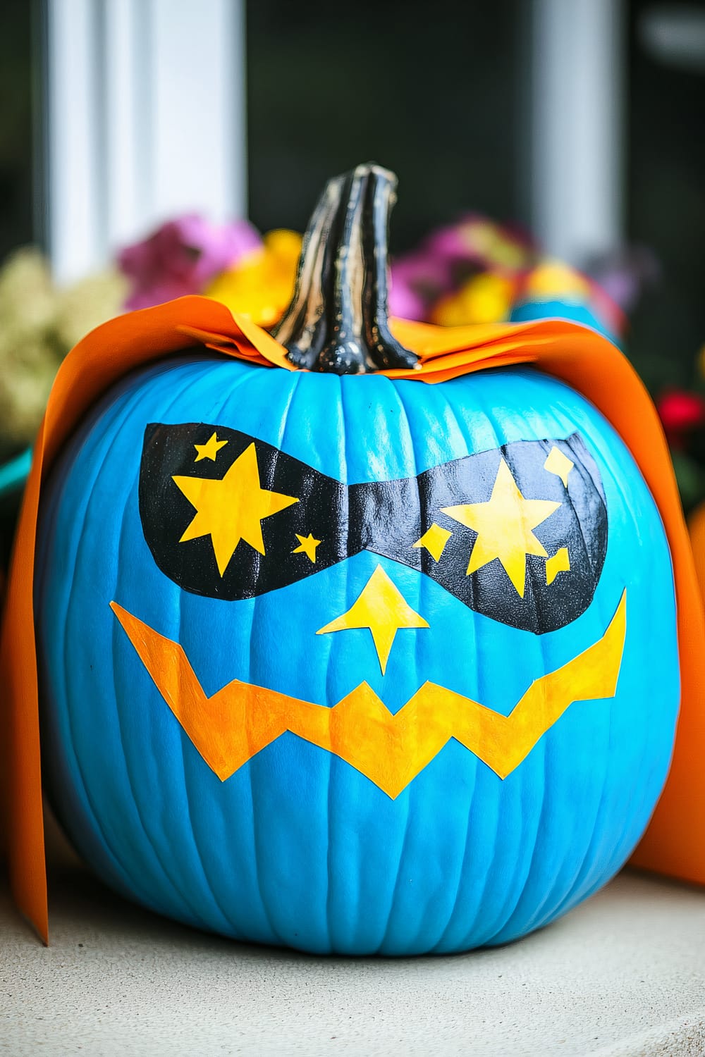 A bright blue pumpkin decorated for Halloween with various vibrant features. The pumpkin has black and yellow starry sunglasses, a yellow star-shaped nose, and a jagged yellow mouth. Additionally, it is adorned with an orange cape on its sides, giving it a superhero-like appearance. In the background, there are some flowers with colors such as yellow and purple, slightly blurred.