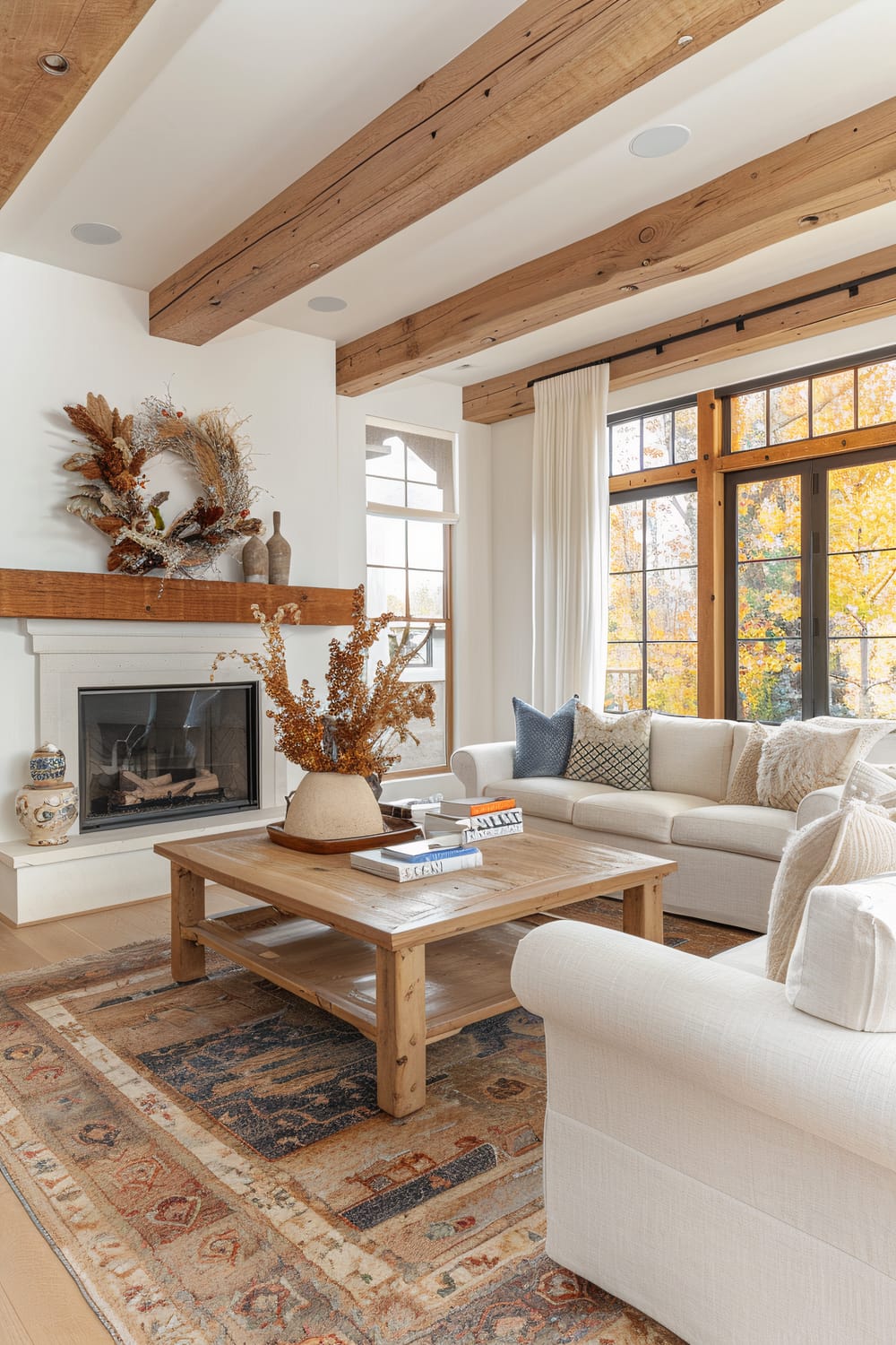 A well-lit living room features a large wooden coffee table with a decorative arrangement of dried plants in a vase. The coffee table sits on a colorful, patterned rug. Surrounding the coffee table are cream-colored sofas adorned with various throw pillows. The backdrop includes large windows with white curtains, a fireplace with a wooden mantel decorated with a seasonal wreath, and two ceramic vases. Exposed wooden beams on the ceiling add a rustic touch to the room.