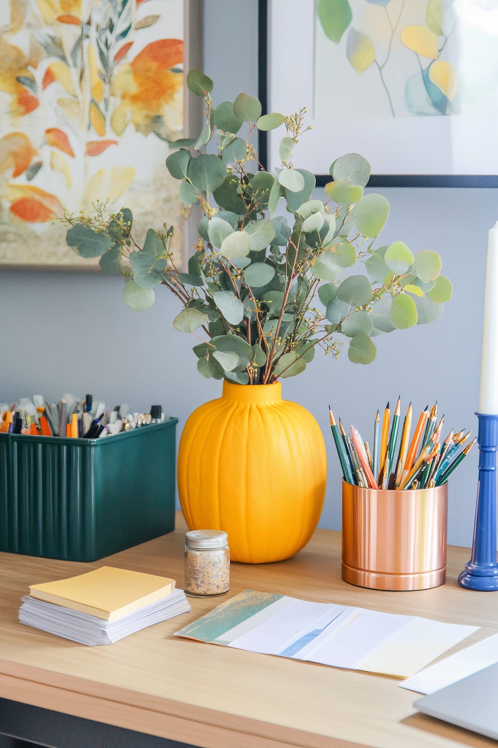 A table set up for a Friendsgiving DIY craft corner, featuring a large yellow vase with eucalyptus branches, a small stack of colored papers, a copper container with colored pencils, a glass jar of decorative flakes, and a variety of craft supplies including scissors and glue sticks. The scene is enhanced by vibrant wall art and natural light from a window, as well as a cobalt blue lamp providing focused lighting.