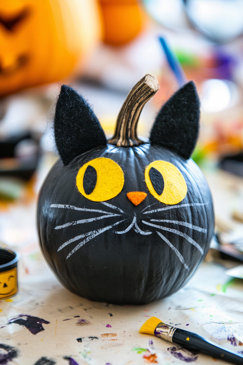 A black pumpkin designed to look like a black cat sits on a table. The pumpkin is painted black with bright yellow felt eyes, an orange felt nose, and white painted whiskers and mouth. Black felt ears are attached to the top of the pumpkin. Surrounding the pumpkin are various art supplies including paintbrushes and jars of paint on a white tablecloth with colorful paint splatters.