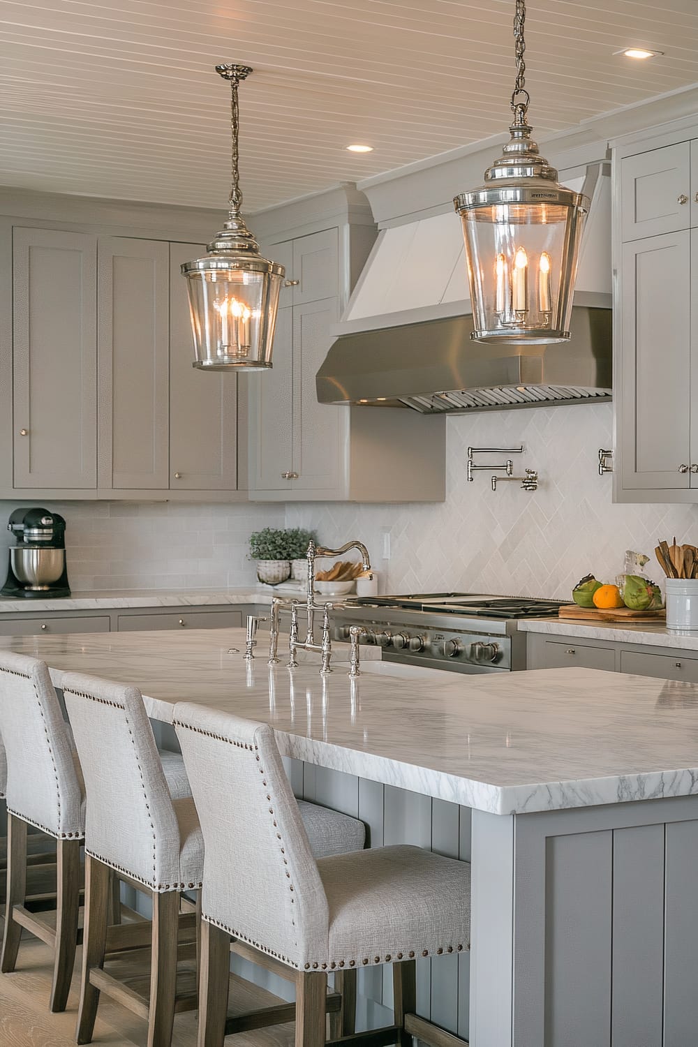 A modern kitchen featuring a marble countertop island with three upholstered bar stools showcasing studded detailing along the edges. Above the island, two large glass pendant lights hang, illuminating the area. The backdrop includes light grey cabinetry, a stainless steel range hood, and a tile backsplash in a herringbone pattern. On the countertop, there is a mixer, a fruit bowl with various fruits, and wooden kitchen utensils in a ceramic holder. The ceiling has recessed lighting and beadboard detailing.