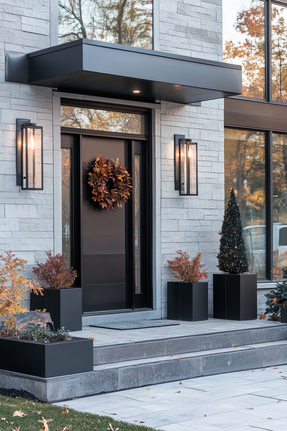 A modern front entrance adorned with seasonal decor. The doorway is black, with a matte finish and has a modern autumn wreath hanging on it. It is framed by two tall glass-paneled windows. The exterior walls are made of light gray bricks, complementing the black door. Large black wall sconces with glass panels are mounted on each side of the door, and they are lit, providing a warm glow. The entrance steps are made of gray stone, leading up to a sleek, dark overhang. Flanking the steps and entrance are minimalistic black planters containing seasonal foliage, and a small festive tree with lights.