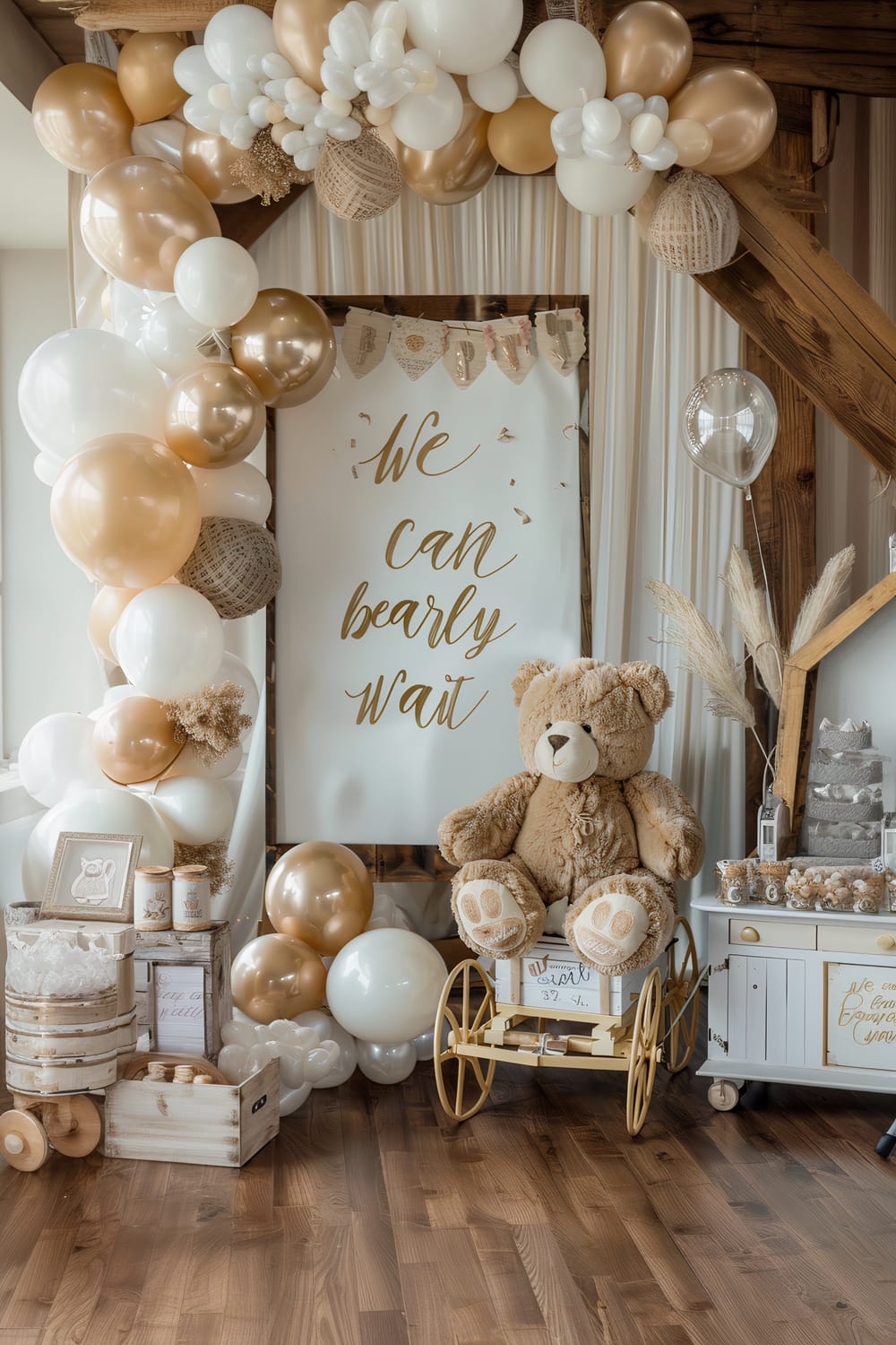 The image depicts a beautifully decorated baby shower area with a soft, rustic theme. Dominating the scene is a large sign that reads "We can bearly wait," surrounded by white and gold balloons arranged in an arch. On the right side, a large teddy bear is seated on a yellow vintage-style cart. To the left, there's a wooden crate with baby items and decorations, including a framed picture, small candles, a bucket with a lace cloth, and a toy wagon. The decor continues with a collection of pampas grass, a grey diaper cake, and more baby-themed items, all placed on a white dresser.