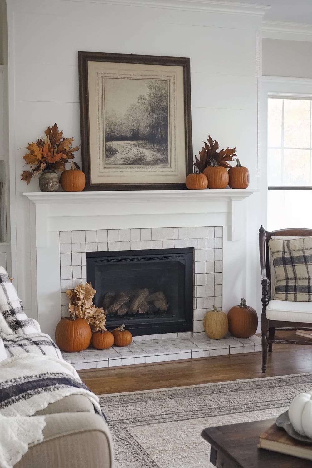 A beautifully arranged living room features a classic white fireplace adorned with small pumpkins and autumn leaves in rustic vases, evoking the essence of fall. Above the mantel rests a large framed black-and-white landscape painting. Next to the fireplace, a traditional armchair with a plaid pillow enhances the cozy atmosphere. A patterned rug in beige and gray tones covers the wooden floor, with a cushy throw blanket draped over the arm of a sofa.