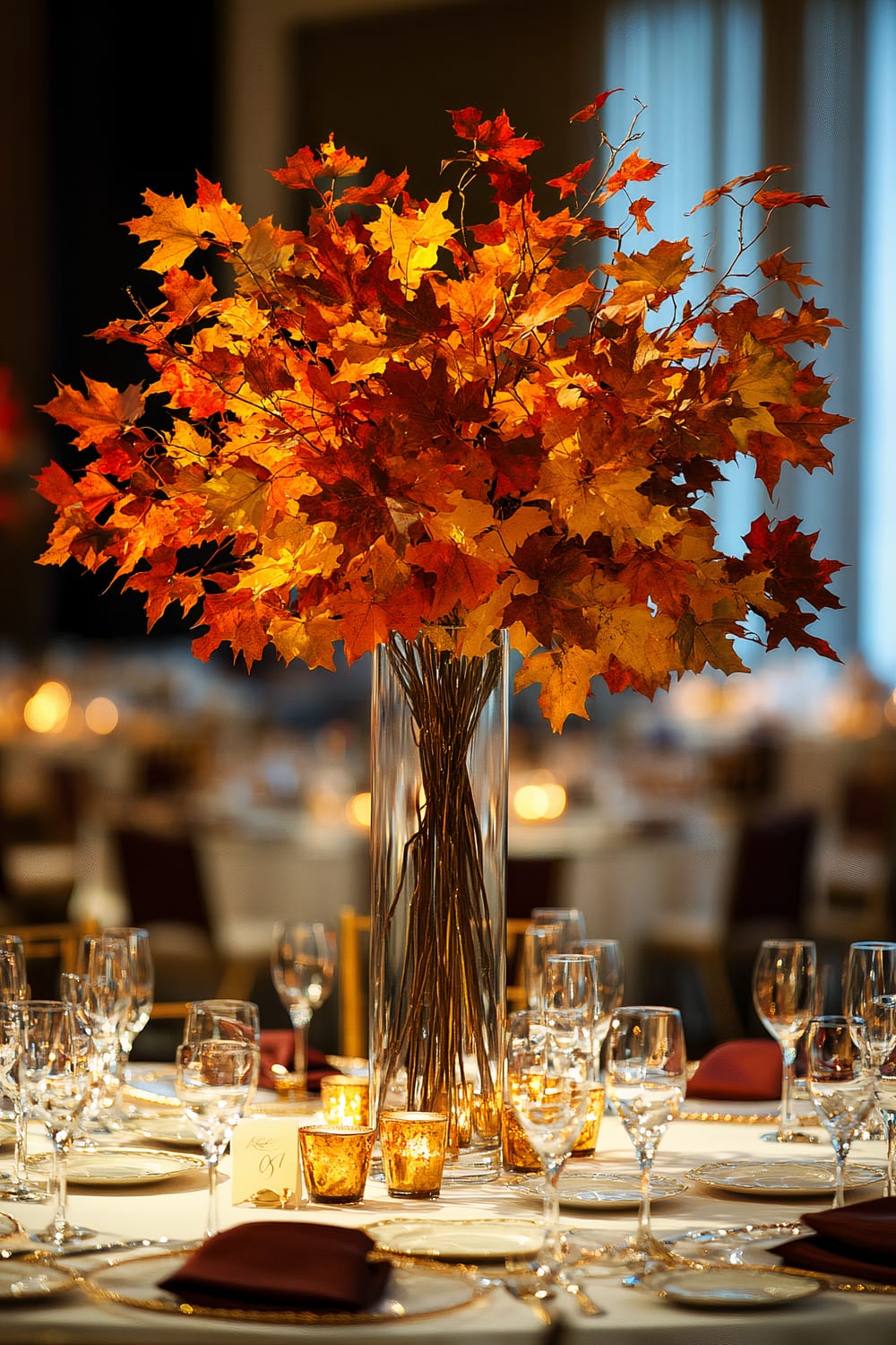 An elegant dining table adorned with a tall centerpiece of autumn leaves in shades of orange and red, enclosed in a glass vase. The table is set with clear glassware, plates, and neatly folded napkins, with small golden candle holders adding a warm glow.