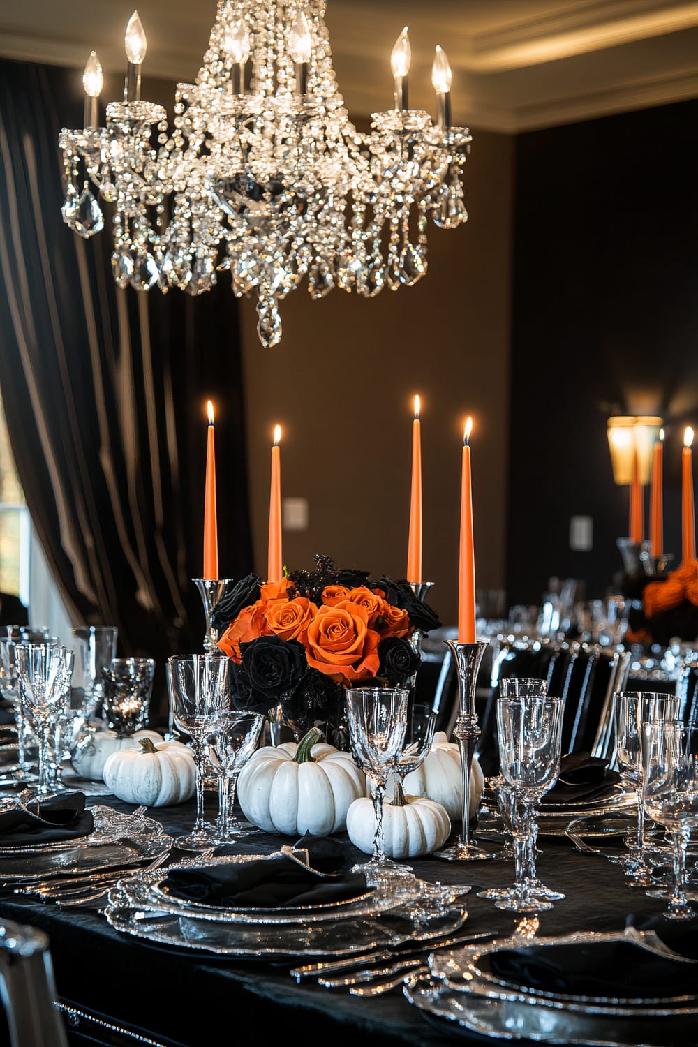 A lavish dining table decorated with Halloween-themed elements, featuring a grand crystal chandelier above. The table is set with elegant glassware and silver tableware on a black tablecloth. Orange taper candles in silver holders along with bouquets of orange and black roses are the focal points, complemented by white pumpkins placed as additional decor.