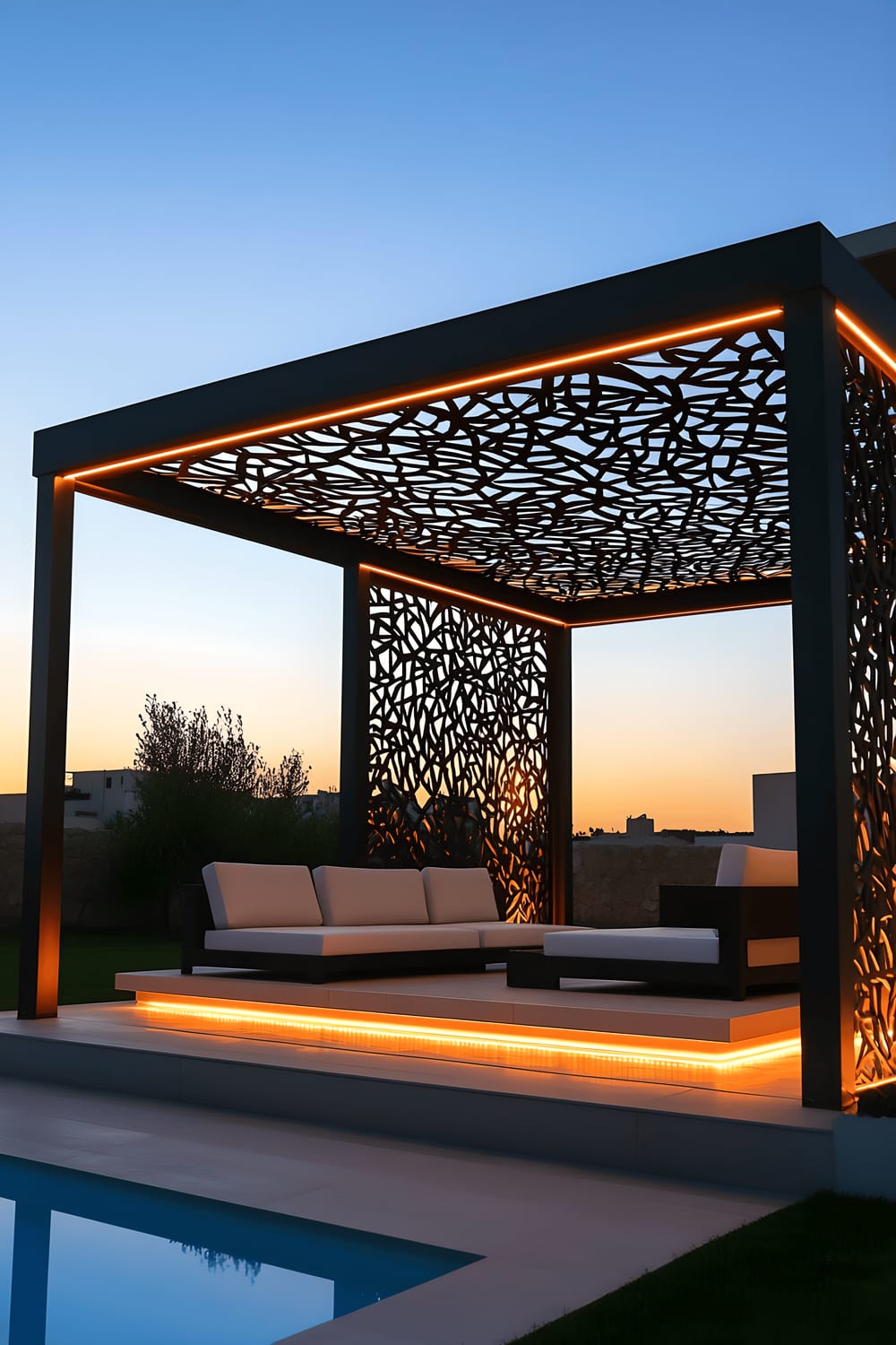 A modern pergola made of sleek black metal beams with intricate geometric patterns lit by integrated LED strip lighting. The pergola houses a minimalist white lounge set with plain cushions, photographed during dusk from a diagonal perspective.