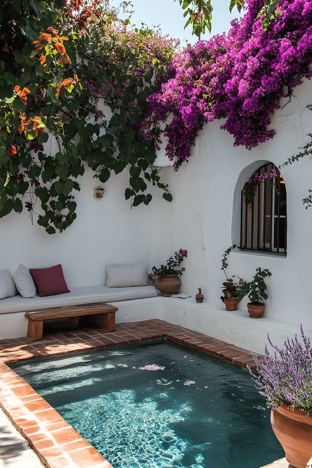 A picturesque Mediterranean courtyard featuring a small plunge pool surrounded by burnished terracotta tiles. The area is framed by white stucco walls adorned with lively bougainvillea blossoms, creating a harmonious blend of organic and built forms. An inviting built-in bench punctuates the vibe of the courtyard, strewn with plush linen cushions in soothing hues. Rustic ceramic planters brimmed with lavender further enrich the sensory experience of the space, weaving an intimate and tranquil atmosphere.