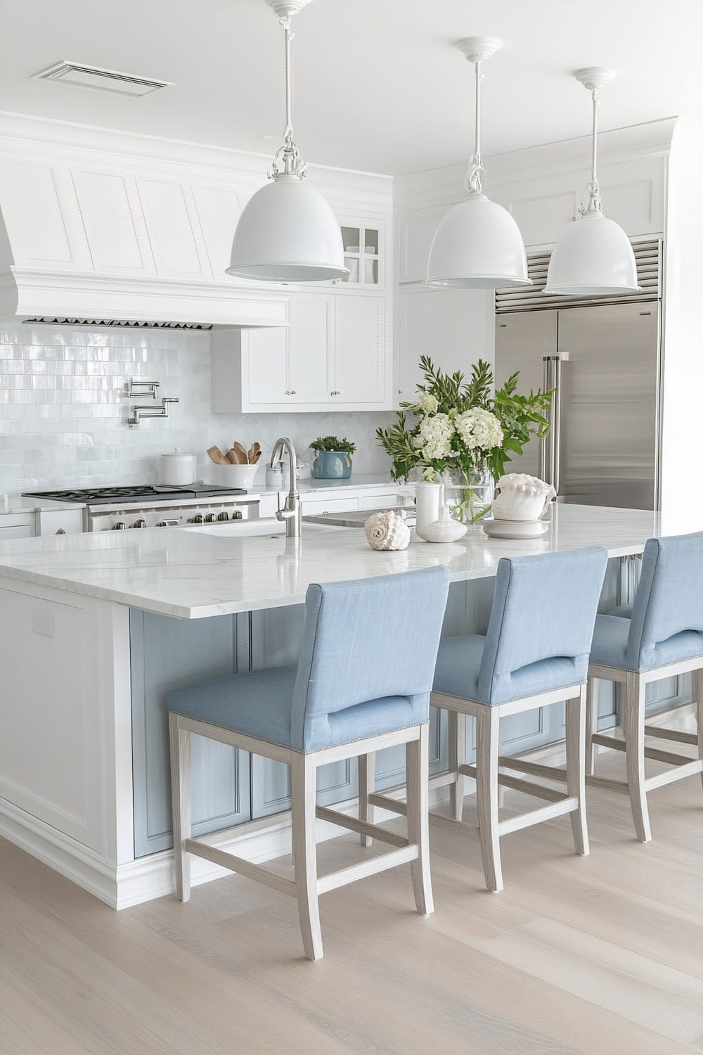 The image showcases a bright and elegant kitchen interior. The kitchen features white cabinetry and a large marble island with a built-in sink. Three light blue cushioned chairs with wooden legs are placed along one side of the island. Overhead, there are three white pendant lights. The backsplash is a glossy white tiled surface, and there is a stainless steel stove with a range hood above it. To the right, a stainless steel refrigerator is visible. Decorative elements, such as a vase of white flowers and various kitchen accessories, are neatly arranged on the island.