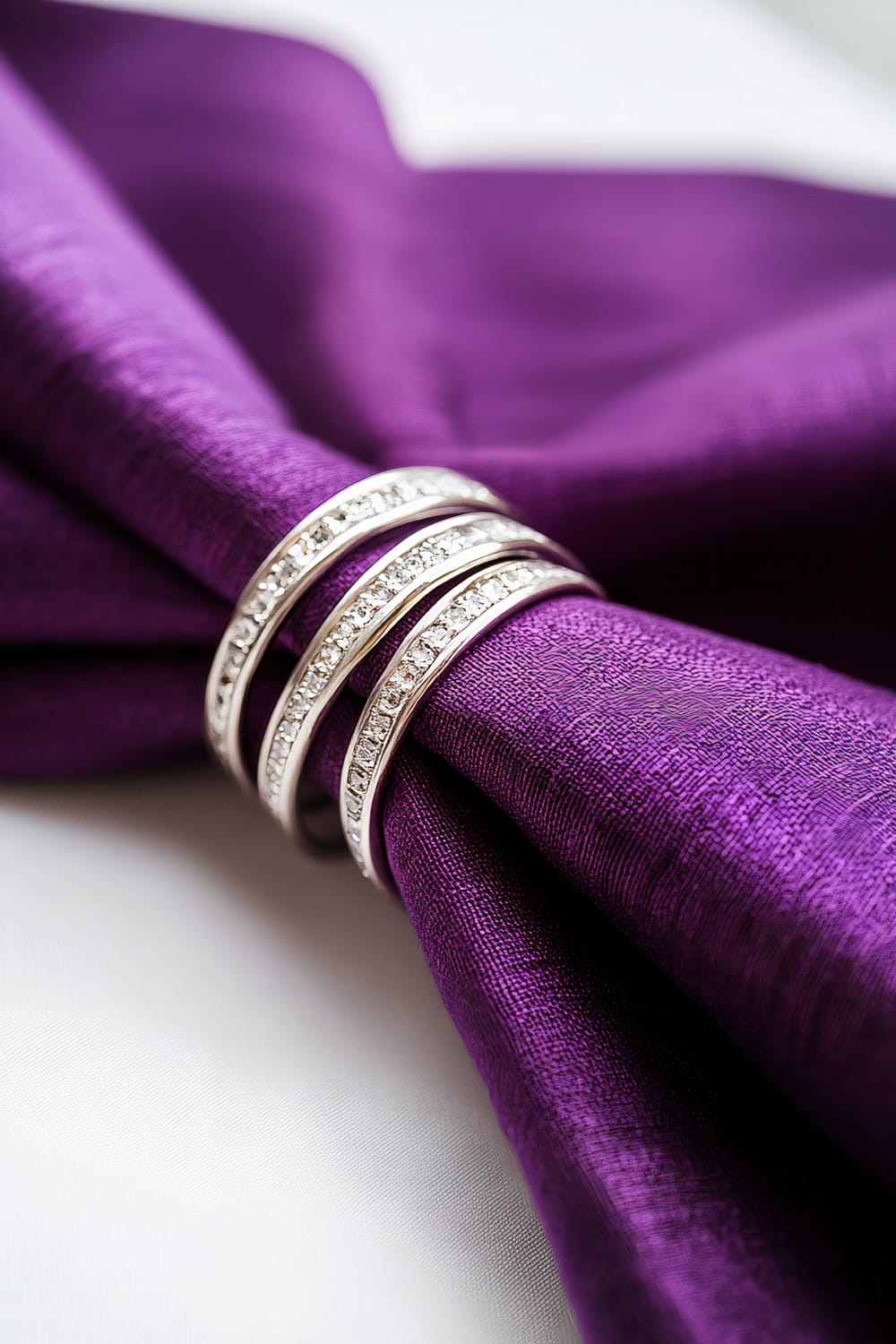 A close-up of deep purple linen napkins neatly folded and held together with elegant silver rings on a stark white kitchen table. Intense top lighting creates a high-contrast effect, highlighting the rich purple hues of the napkins and the metallic sheen of the rings.