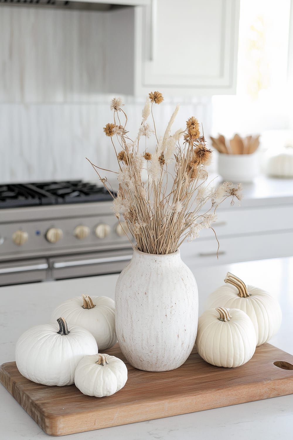 A minimalist kitchen countertop display featuring a large ceramic vase with dried grasses and flowers, positioned on a wooden cutting board. Surrounding the vase are several small white pumpkins. In the background, a modern stove with light-colored cabinetry and additional white pumpkins and a bowl of utensils are visible.