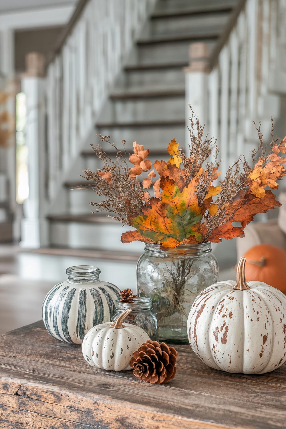 An autumnal indoor decor scene showing a wooden table adorned with two small white decorative pumpkins, a small candle holder, a pinecone, and a jar filled with dried autumn leaves and branches. The background features a rustic staircase with a white railing.