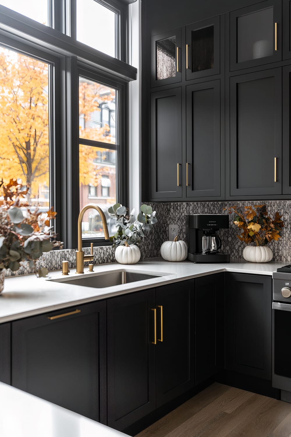 A contemporary kitchen featuring elegantly dark cabinetry with sleek, golden handles. A white marble countertop hosts seasonal white pumpkins and eucalyptus arrangements, adding a touch of autumn decor. The stylish backsplash displays a geometric pattern, pairing harmoniously with the minimalist stainless steel appliances and a modern, gold faucet. Large windows flood the space with natural light, illuminating the interior and offering a scenic view of colorful fall foliage outside.