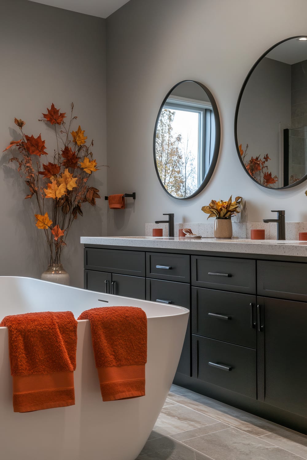 A modern bathroom features a freestanding white bathtub adorned with two bright orange towels, contrasting against the neutral gray walls. The vanity set with twin sinks showcases a minimalist gray countertop, under which sits a row of sleek black cabinetry with silver handles. Mounted above the sinks are two large, round mirrors reflecting a bright scene outside the window. Autumnal decorations, including vibrant orange and yellow leaves in vases, accentuate the seasonal theme, lending warmth to the contemporary design.