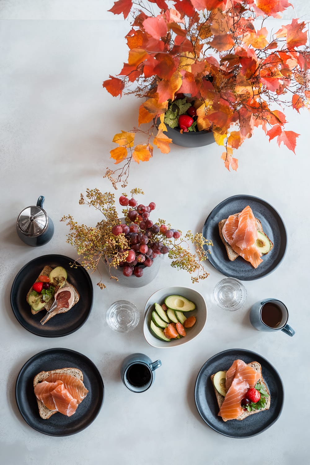 A minimalist brunch setting on a white marble table with earth-toned ceramic plates holding avocado toast topped with smoked salmon and fresh fruit. In the center is a deep yellow geometric metallic centerpiece with red leaves and autumn branches. There's a small metallic vase with fresh greenery and five cobalt blue glass mugs filled with herbal tea. The table is illuminated with striking lighting against a white backdrop.