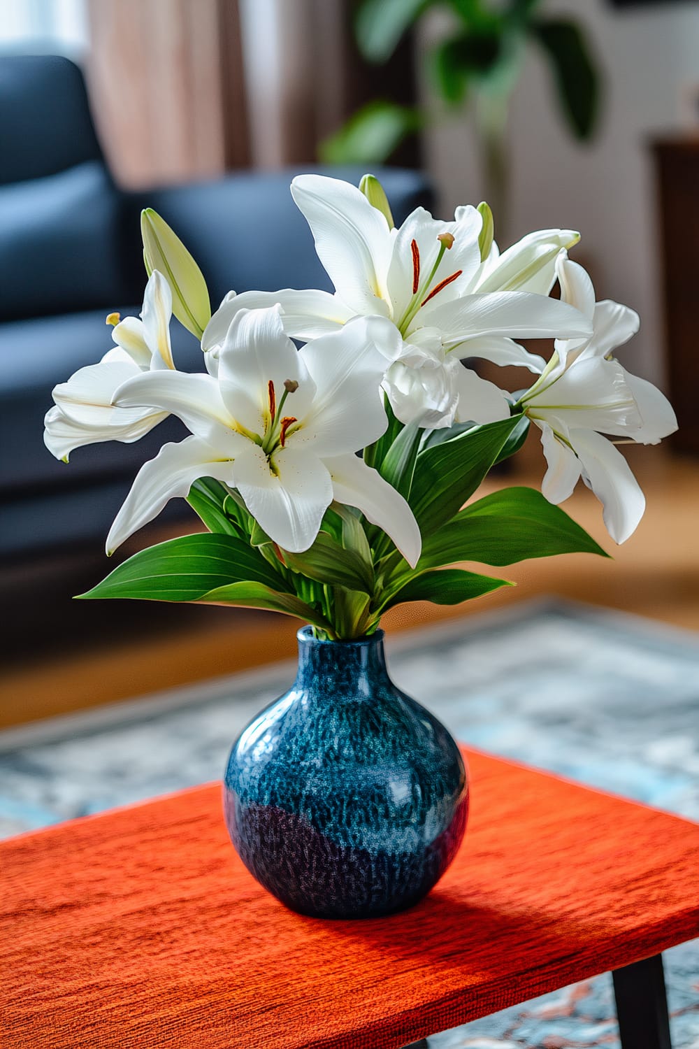 A contemporary coffee table with a vibrant orange runner, featuring a blue vase holding an arrangement of white lilies with green leaves. The background includes blurred elements of a dark-toned sofa, soft ambient lighting, and partial views of houseplants and furniture.