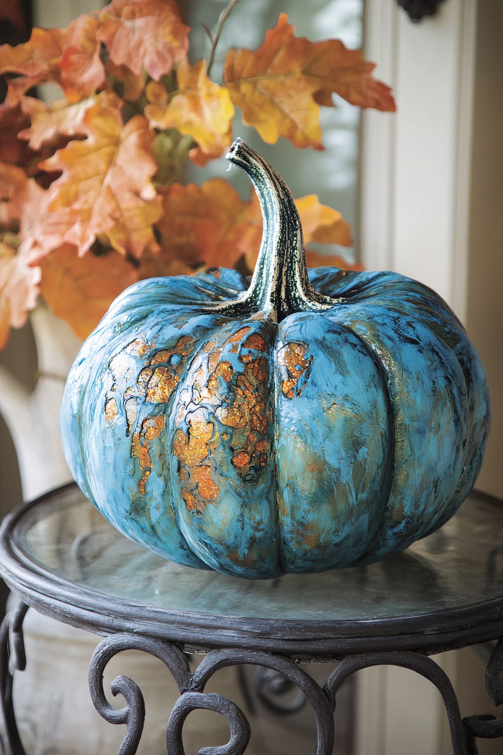 A close-up image of a decorative pumpkin painted in a vibrant blue color with gold and copper-colored accents, giving it a textured and artistic look. The pumpkin is placed on a glass tabletop with ornate wrought iron legs. In the background, there are autumn leaves in shades of orange, yellow, and brown, creating a seasonal ambiance.