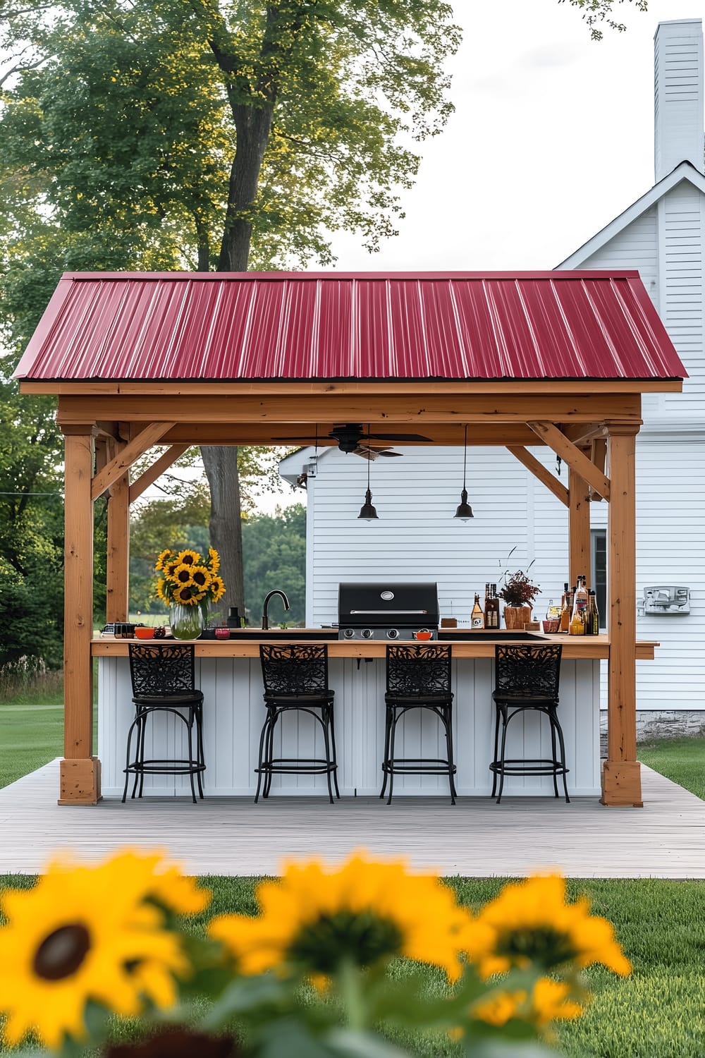 A backyard setup features a wooden gazebo with a charming red metal roof. Underneath, a built-in bar environment encircles a grill, with counters for seating on sleek black barstools. A vase of bright sunflowers and casual dining ware accent the area, adding a touch of homeliness. In the background, a white house with a wooden deck is visible.