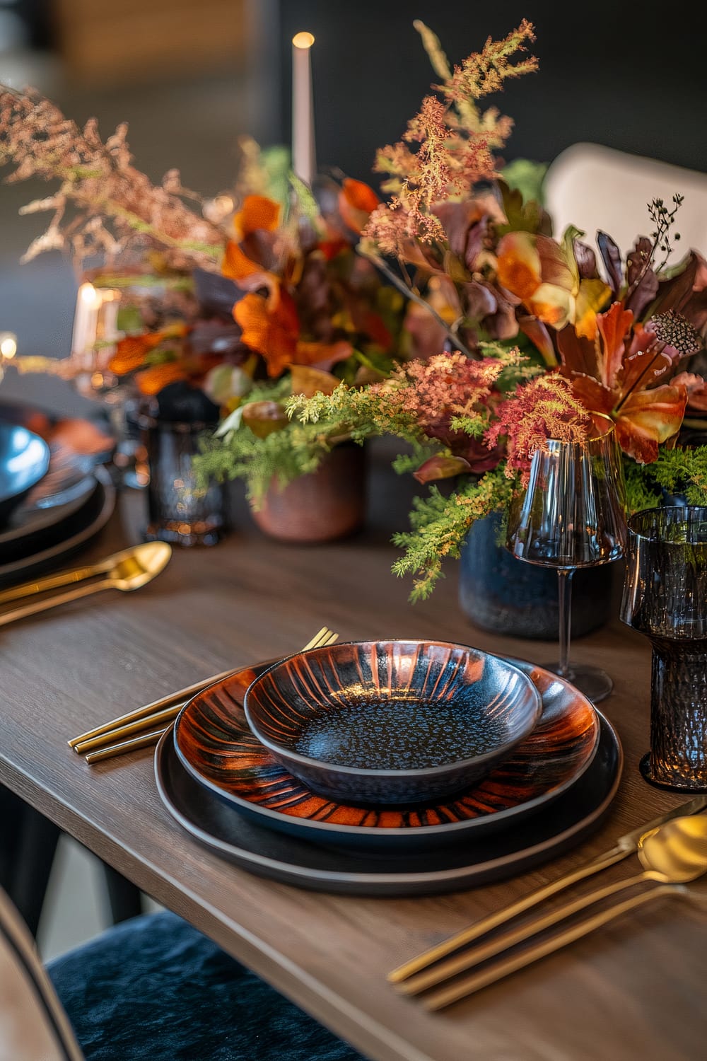 A modern Thanksgiving table setting featuring deep navy and burnt orange accents. The table includes geometric ceramic plates with a speckled pattern, gold flatware, and vibrant potted centerpieces with autumnal foliage. The ambient lighting casts soft, dramatic shadows, creating an elegant yet inviting atmosphere.