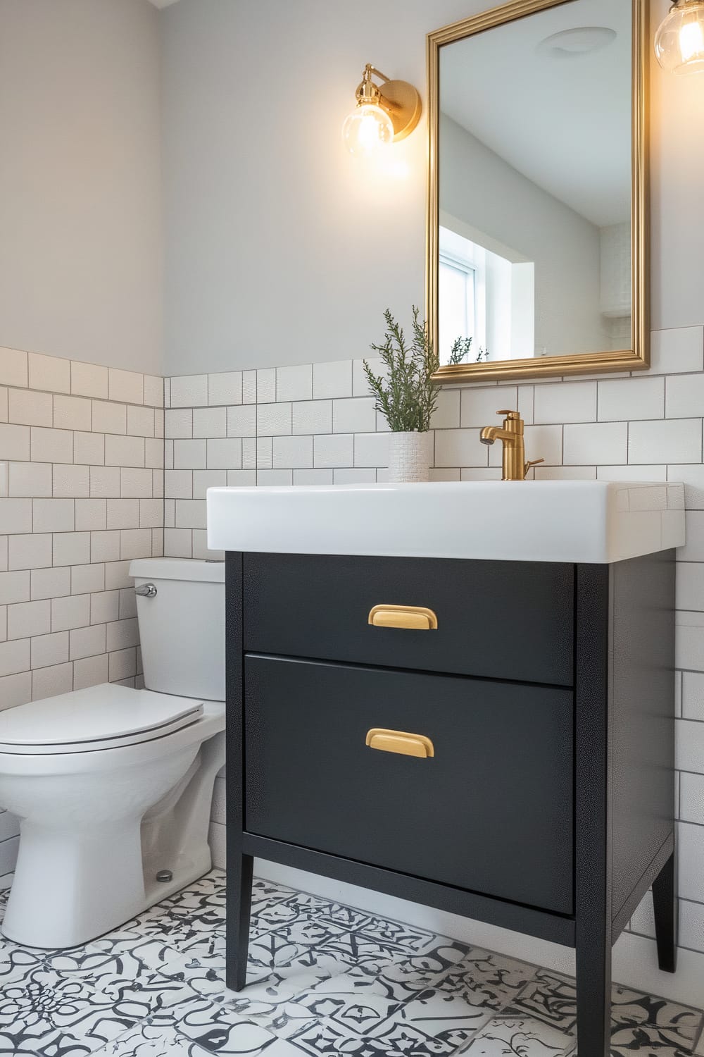 A small bathroom featuring a sleek black vanity with brass handles and a white countertop. Above the vanity is a rectangular mirror framed in brass, illuminated by brass wall sconces with exposed bulbs on either side. The walls are partially tiled with white subway tiles, and the floor features a bold, black-and-white patterned tile. A small potted plant sits on the vanity, adding a touch of greenery.