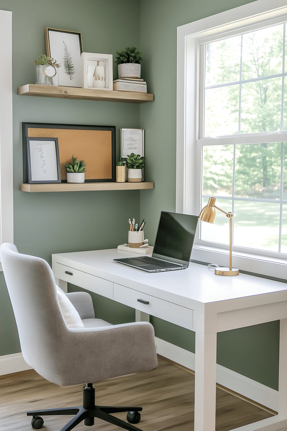 A contemporary home office in Milan with a sleek white desk and a high-backed ergonomic chair in charcoal gray. Above the desk are floating wooden shelves containing neatly arranged books, a brass desk lamp, and small terrariums with succulents. The backdrop is a soft sage green wall, and a minimalist black-framed corkboard displaying inspirational quotes and pinned photos hangs to the side. The space is brightened by large windows