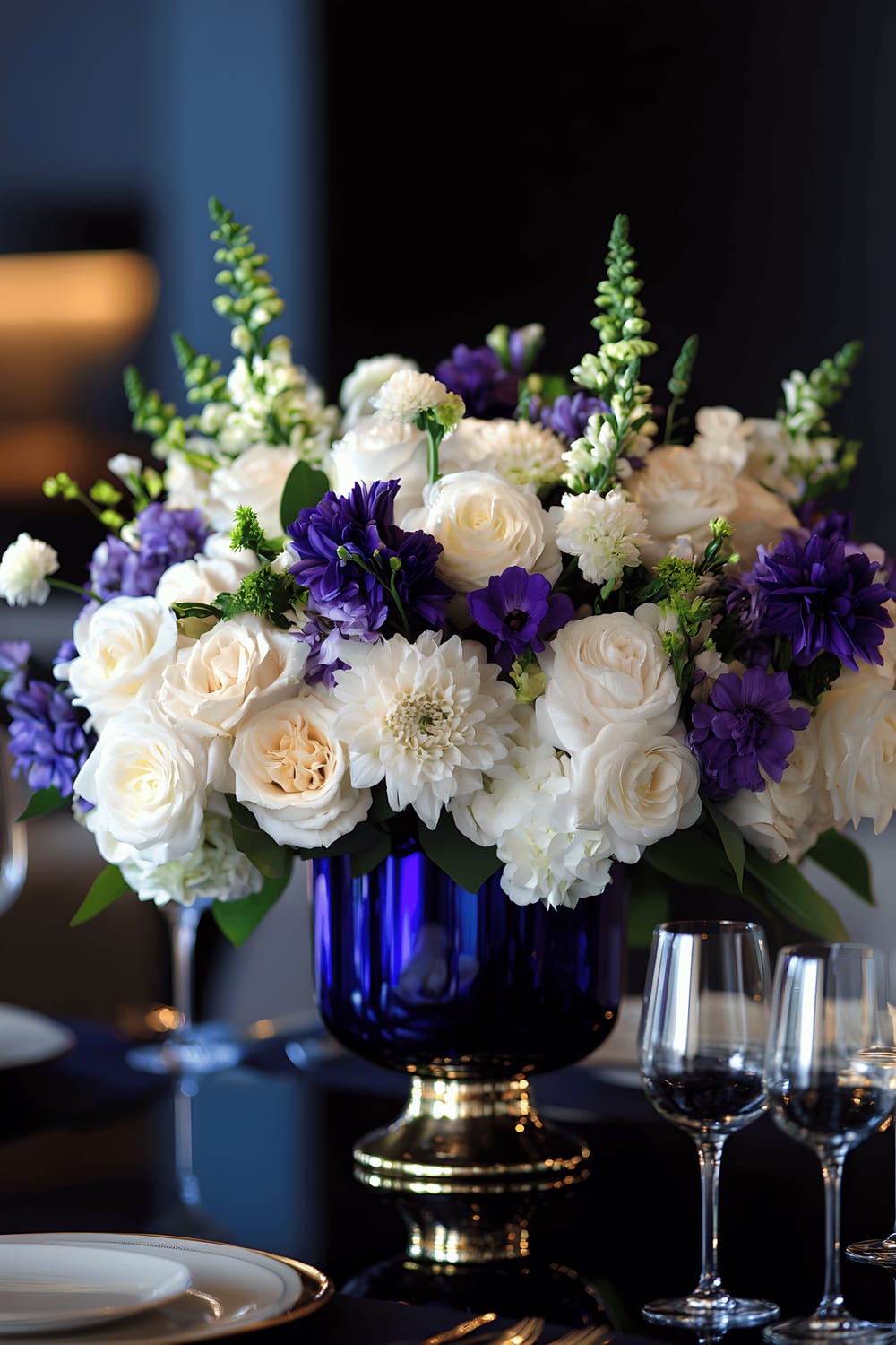 An elegant dining room featuring a centerpiece on a dark wood table. The centerpiece includes a cascade of white and purple flowers arranged on a large cobalt blue brass mirror base, reflecting the surroundings. This is set against a minimalist backdrop of dark walls and modern furniture, and complemented by soft, directional lighting.