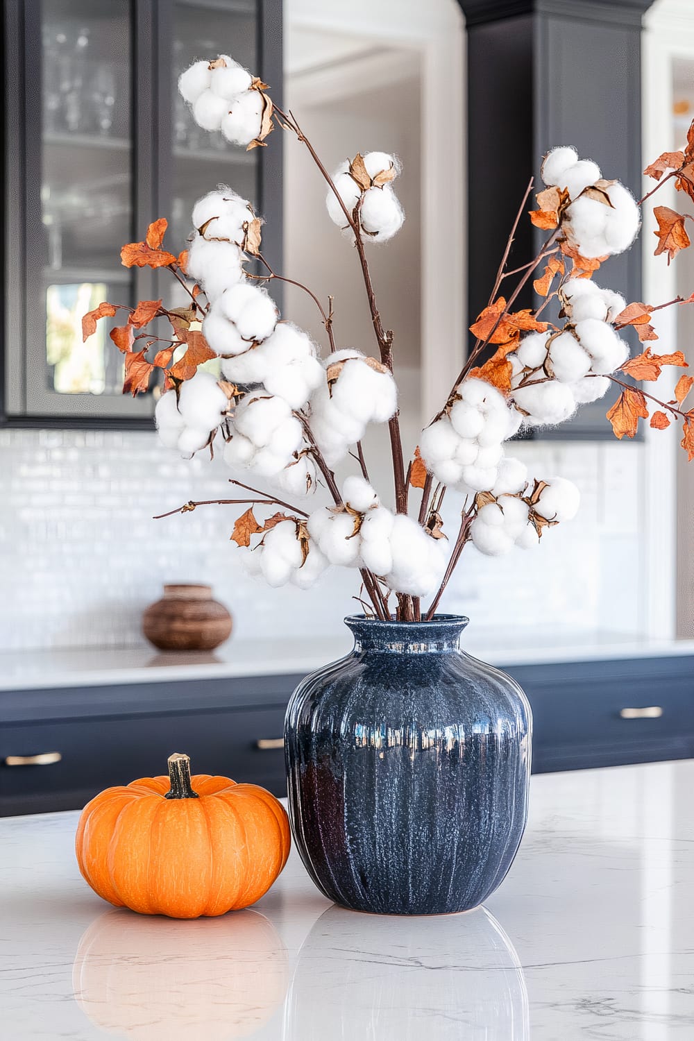 A navy blue vase filled with cotton branches and autumnal leaves sits on a white marble countertop. Next to the vase is a small orange pumpkin. In the background, there are dark cabinetry with glass doors revealing glassware, a white tiled backsplash, and another decorative piece on a counter.