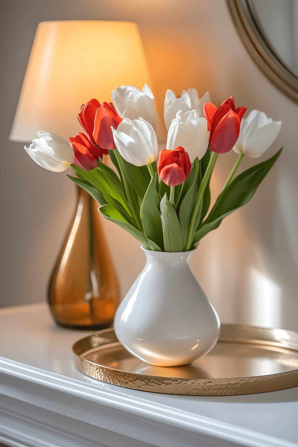 A sleek white porcelain vase filled with a balanced arrangement of red and white tulips residing on a metallic gold tray which is placed on a light gray mantel. The scene is bathed in the warm light from an amber-colored lamp nearby, reflecting off the gold tray and enhancing the vivacity of the tulip colors.